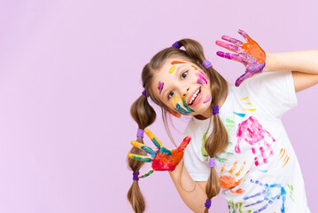 A beautiful little girl stained in multicolored paints on a pink isolated background has fun...