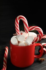 Cup of tasty cocoa with marshmallows and Christmas candy canes on black wooden table