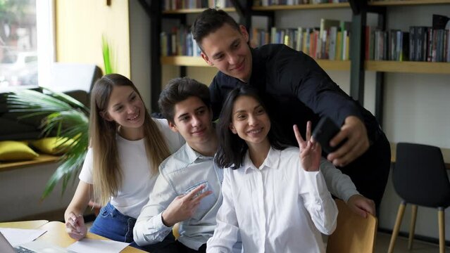European students, group of four take selfie in the college or university library. Group of four standing in front bookshelves, happily smiling, gesturing, showing peace gesture