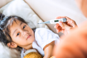 Obraz na płótnie Canvas Sick kid. Mother checking temperature of her sick daughter with thermometer in mouth, child laying in bed taking measuring her temperature for fever and illness