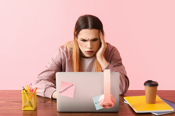 Displeased transgender programmer with laptop at table on pink background