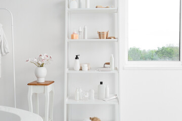 Fototapeta na wymiar Shelf unit with different bath accessories and vase with flowers on table near white wall