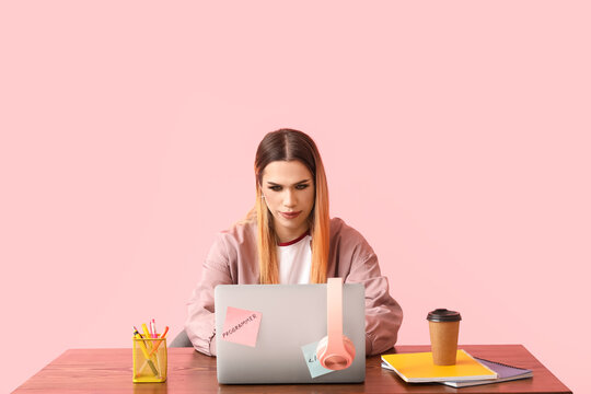 Transgender programmer with laptop at table on pink background