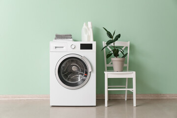 Washing machine with detergent, towels and houseplant on chair near green wall