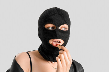 Young woman in balaclava applying red lipstick against light background