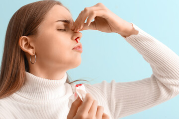 Young woman with nosebleed and tissue on blue background, closeup