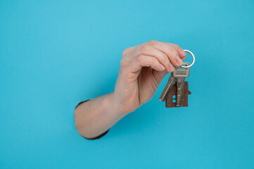 A woman's hand holds the keys with a keychain in the shape of a house on a blue background. 