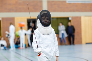 Fototapeta na wymiar Little kid boy fencing on a fence competition. Child in white fencer uniform with mask and sabre. Active kid training with teacher and children. Healthy sports and leisure.