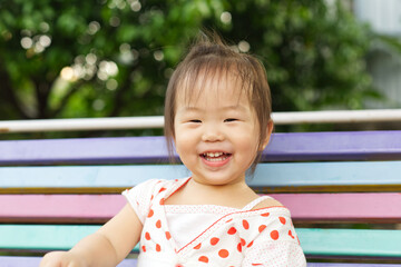 A happy Asian toddler is sitting on colorful chair with blur green trees in background from Thailand.