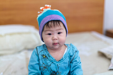 A cute Asian baby is sitting on the bed with colorful hat and blue cloth.  An Asian baby with red spots from mosquito bites on her face.