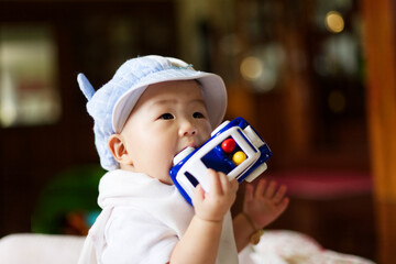 A cute Asian baby with a blue hat is putting a car toy into her mouth.