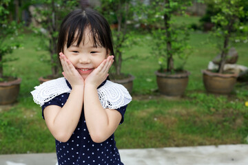 An Asian cute girl in action of 2 hands on her cheek with background of green trees in the pots and copy space from Thailand.