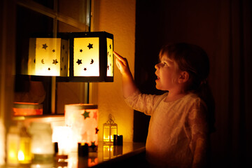 Little kid girl sitting by window with selfmade hand crafted lanterns with candles for St. Martin...