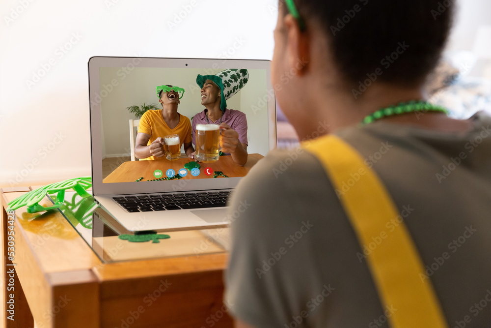 Poster Mixed race woman making st patrick's day video call to friends holding beers on laptop at home