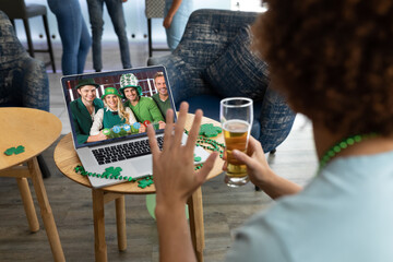Mixed race man at bar holding beer making st patrick's day video call waving to friends on laptop