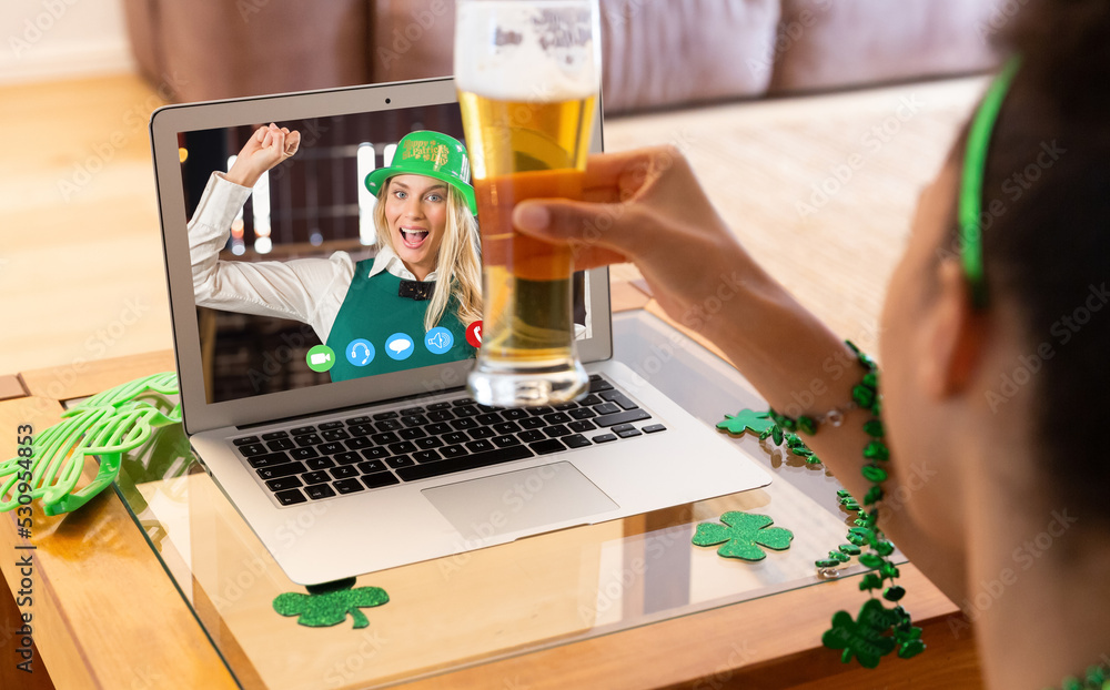 Sticker Mixed race woman with beer having st patrick's day video call with female friend on laptop at home