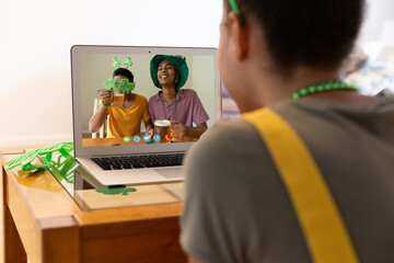 Mixed race woman making st patrick's day video call to male friends holding beers on laptop at home - Powered by Adobe