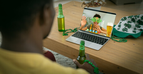 African american man holding beer having st patrick's day video call with friend on laptop at home