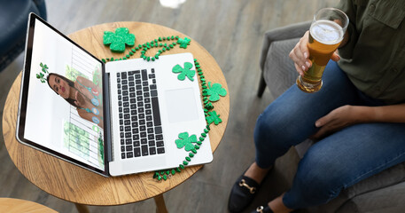 Mixed race woman holding beer at bar making st patrick's day video call with friend on laptop