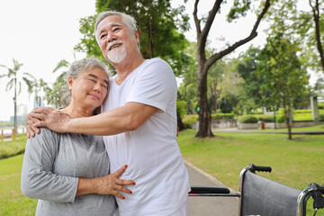 Obraz premium Asian senior woman or caregiver helping senior man walk with wheelchair at park outdoor. Elderly wife taking good help care and support of elder husband patient outside the retirement house.