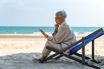 Modern Asian senior businesswoman working on digital tablet with internet at tropical beach in sunny day. Elderly retired woman enjoy outdoor lifestyle with using technology on summer holiday vacation