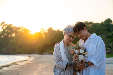 Happy Asian family senior couple enjoy outdoor lifestyle on summer beach vacation. Elderly husband surprise his wife with flower bouquet for celebrating marriage anniversary on the beach at sunset.