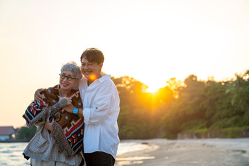 Fototapeta premium Happy Asian family senior couple enjoy outdoor lifestyle at the beach on summer holiday vacation. Senior man husband cover his wife with a blanket and hugging together on the beach at summer sunset