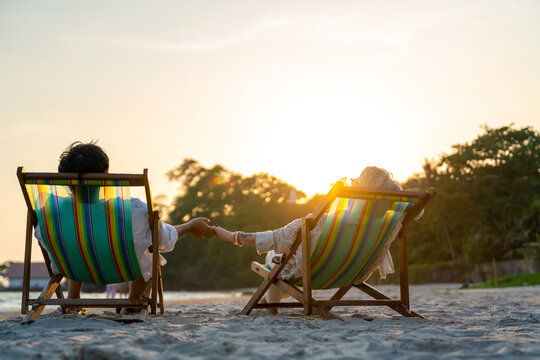 Happy Asian family senior couple enjoy outdoor lifestyle on summer beach holiday vacation at the sea. Elderly retired man and woman relaxing on beach chair and holding hands together at summer sunset