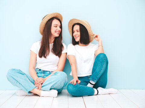 Two young beautiful smiling hipster female in trendy same summer white t-shirt and jeans clothes. carefree women posing near blue wall in studio. Cheerful and positive models in hats