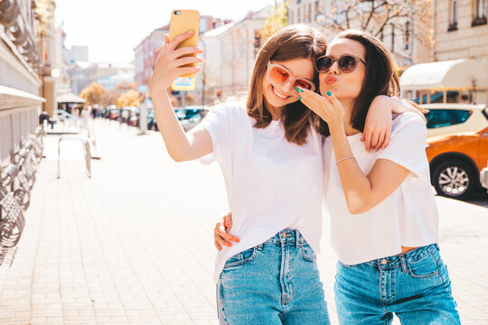 Two young beautiful smiling hipster female in trendy summer white t-shirt clothes and jeans.Sexy carefree women posing on the street background. Positive models having fun. Taking selfie photos