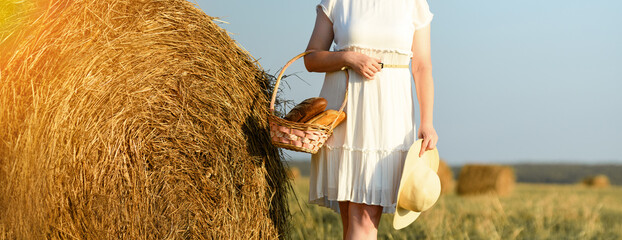 Girl and bread. A woman in a white dress stands with a basket of bread against the backdrop of a haystack and the sky. Harvest concept. Banner