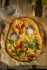 Garden art focaccia bread with vegetables, greens, herbs - beautiful flower composition on homemade freshly baked bread, salty vegan pastry. Wooden table. Copy space. Italian cuisine. Top view