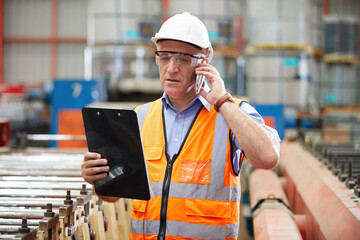 male factory workers or engineer talking on smartphone and looking information on clipboard in...