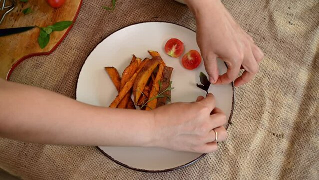 View from above of a chef's hand serving a dish with roasted sweet potatoes with halves of cherry tomatoes and rosemary leaves. Healthy vegan meal. Baked wedges of organic batata.