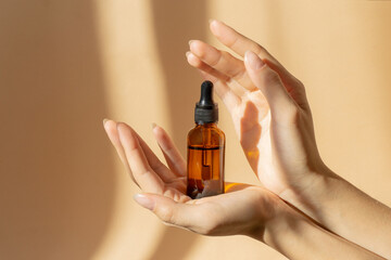 Women's hands hold bottle of amber glass with cosmetic serum on brown background. Mockup of...