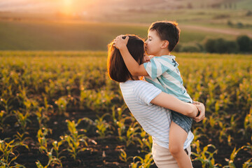 Happy boy kissing his mother on the forehead standing in their cornfield at sunset. They are...