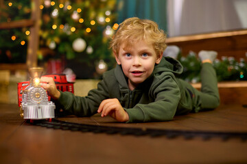 Fototapeta na wymiar Cute curly-haired boy is playing with a magic toy train at the Christmas tree. Concept of celebrating Christmas and New Year.