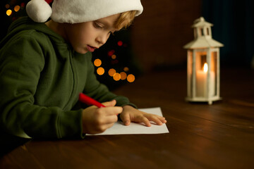 Boy in a Christmas hat writes a letter to Santa Claus by the light of a lantern. Child writes a...