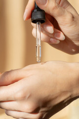 Close-up of pipette with collagen moisturizing hyaluronic serum in woman's hand in rays of sunlight. Vertical image beige background