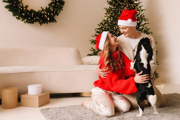 A couple in love, husband and wife in red Santa Claus hats, play with pet and celebrate the Christmas holiday in decorated living room with a tree in winter. The couple is preparing for the new year 