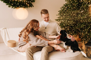 A young man and a woman in cozy sweaters are hugging together laughing and playing with a pet dog sitting on a sofa in a decorated interior on a Christmas holiday in the living room of the house