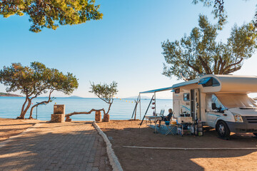 Family traveling with motorhome are eating breakfast on a beach. Travelers on an active family vacation with motorhome RV parked on the beach under a tree facing the sea, Crete, Greece. - obrazy, fototapety, plakaty