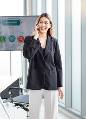 Millennial Asian happy successful professional female businesswoman in formal suit standing holding smartphone smiling talking with customer in company office meeting room with presentation monitor