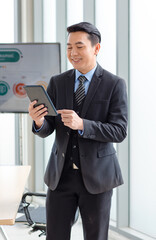 Millennial Asian happy successful professional male businessman entrepreneur in formal suit standing holding touchscreen tablet computer smiling in company office meeting room with big screen monitor