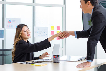 Millennial Asian successful professional female businesswoman in formal suit sitting at working desk in company office meeting room shaking hands smiling greeting with male businessman colleague