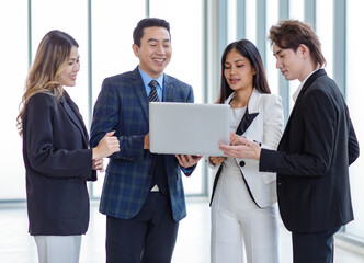 Millennial Asian successful professional businessman manager mentor in formal suit standing holding laptop computer talking helping advising male and female businesswomen colleagues in company office