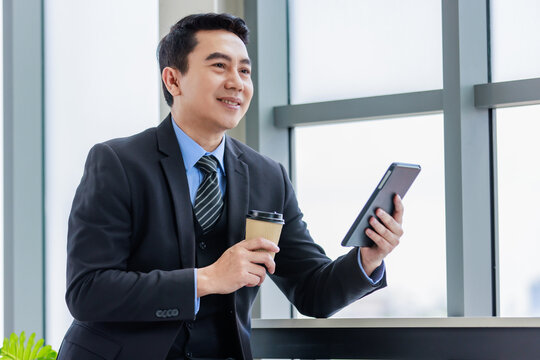 Millennial Asian successful professional male businessman manager in formal suit take break sit smiling holding drinking hot coffee from disposable paper cup and browsing internet online via tablet - Powered by Adobe