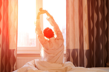 Back view caucasian woman stretching arms and body near window after waking up in bedroom at home. Concept for starting a new day with happiness. Young happy housewife in white shirt