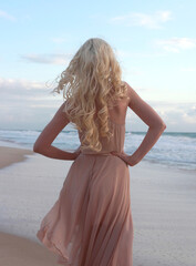 Fototapeta na wymiar full length portrait of beautiful young woman with long hair wearing flowing dress, standing pose walking away from the camera. ocean beach background with sunset lighting.