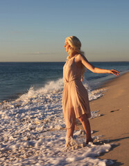 full length portrait of beautiful young woman with long hair wearing flowing dress, standing pose with gestural arms movement .  ocean beach background with back lit sunset lighting.